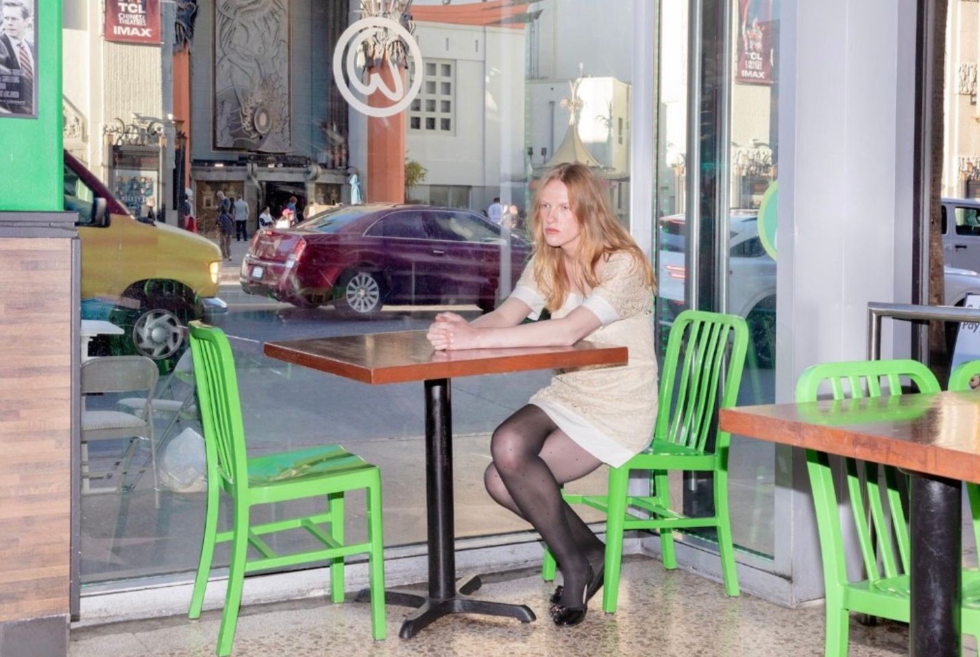 A woman with blonde hair wearing a brown dress and black tights and shoes sits at a table a table alone by a window